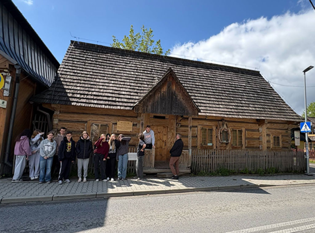 Grupa uczniów stoi na chodniku przed drewnianym budynkiem Muzeum Powstania Chochołowskiego w Chochołowie. Budynek ma drewniane ściany i dach kryty gontem, z małym płotkiem od frontu. Nad wejściem znajduje się tabliczka z napisem „Muzeum”. Obok widać znak przejścia dla pieszych, fragment ulicy oraz niebieskie niebo z białymi chmurami. Uczniowie stoją w grupie, część patrzy w stronę aparatu, a w tle panuje spokojna, wiejska atmosfera.