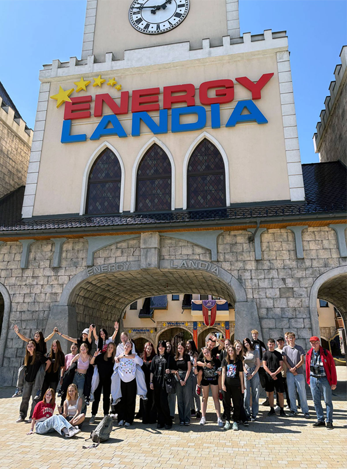 Grupa uczniów stoi i siedzi przed wejściem do parku rozrywki Energylandia w Zatorze. Budynek wejściowy ma jasną fasadę przypominającą zamek z wieżą zegarową na górze. Nad trzema łukowatymi oknami widnieje duży napis „ENERGYLANDIA”. Uczniowie stoją na placu wyłożonym jasną kostką brukową, część osób siedzi z przodu, reszta stoi z tyłu, niektórzy mają ręce uniesione w górę, a inni uśmiechają się i pozują do zdjęcia. W tle pod łukami widać wejścia z napisem „Wejście” oraz kolorowe flagi. Dzień jest słoneczny, a niebo bezchmurne, co nadaje zdjęciu radosny charakter.