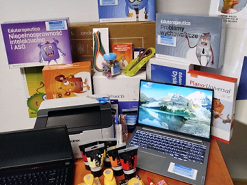 Na zdjęciu widoczny jest sprzęt i materiały dydaktyczne ustawione na stole i w tle przy ścianie. Widać dwa laptopy (jeden otwarty, pokazujący ekran z górami i godziną 09:39, drugi zamknięty), drukarkę Brother, kolorowe farby plakatowe w tubkach, kleje w sztyfcie, markery oraz słoik z kolorowymi spinaczami. W tle znajdują się pudełka z pomocami edukacyjnymi m.in. „Eduterapeutica Niepełnosprawność intelektualna i ASD”, „Eduterapeutica Problemy wychowawcze”, „Eduterapeutica Dysleksja”, „Plano Universal” oraz zestawy gier i materiałów edukacyjnych z rysunkami kolorowych postaci. Wszystko ułożone jest w sposób estetyczny, pokazując przygotowanie materiałów do pracy z uczniami w szkole.