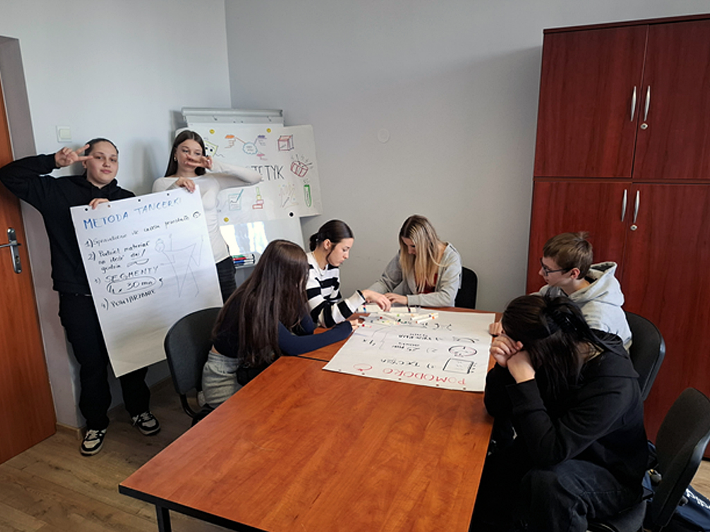 Na zdjęciu widać grupę uczniów podczas zajęć warsztatowych w sali lekcyjnej. Po lewej stronie dwie uczennice stoją i prezentują plakat zatytułowany „METODA TANCERKI”, na którym widnieje krótka instrukcja oraz rysunek postaci. Jedna z uczennic pokazuje znak „V” palcami, a druga uśmiecha się, również pokazując znak pokoju. Po prawej stronie przy stole siedzi pięcioro uczniów, którzy wspólnie pracują nad dużym arkuszem papieru z napisem „Pomodoro”. Uczniowie rysują i zapisują treści markerami, angażując się w tworzenie plakatu projektowego. Jedna z uczennic siedzi z głową opartą na rękach, jakby  zastanawiała się nad kolejnymi krokami. W tle stoi tablica suchościeralna z kolorowymi rysunkami i napisami, obok stoi zamknięta szafa w kolorze ciemnego drewna. Na stole leżą markery i inne przybory potrzebne do pracy w grupach. Całość przedstawia aktywną pracę projektową podczas zajęć edukacyjnych w luźnej, przyjaznej atmosferze.