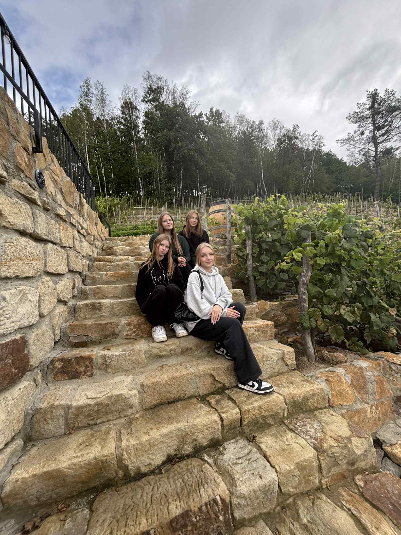 cztery te same dziewczyny stoją razem w pomieszczeniu winnicy Talaska w piwniczce z winami. Za nimi znajduje się drewniany regał wypełniony butelkami wina ustawionymi na półkach.