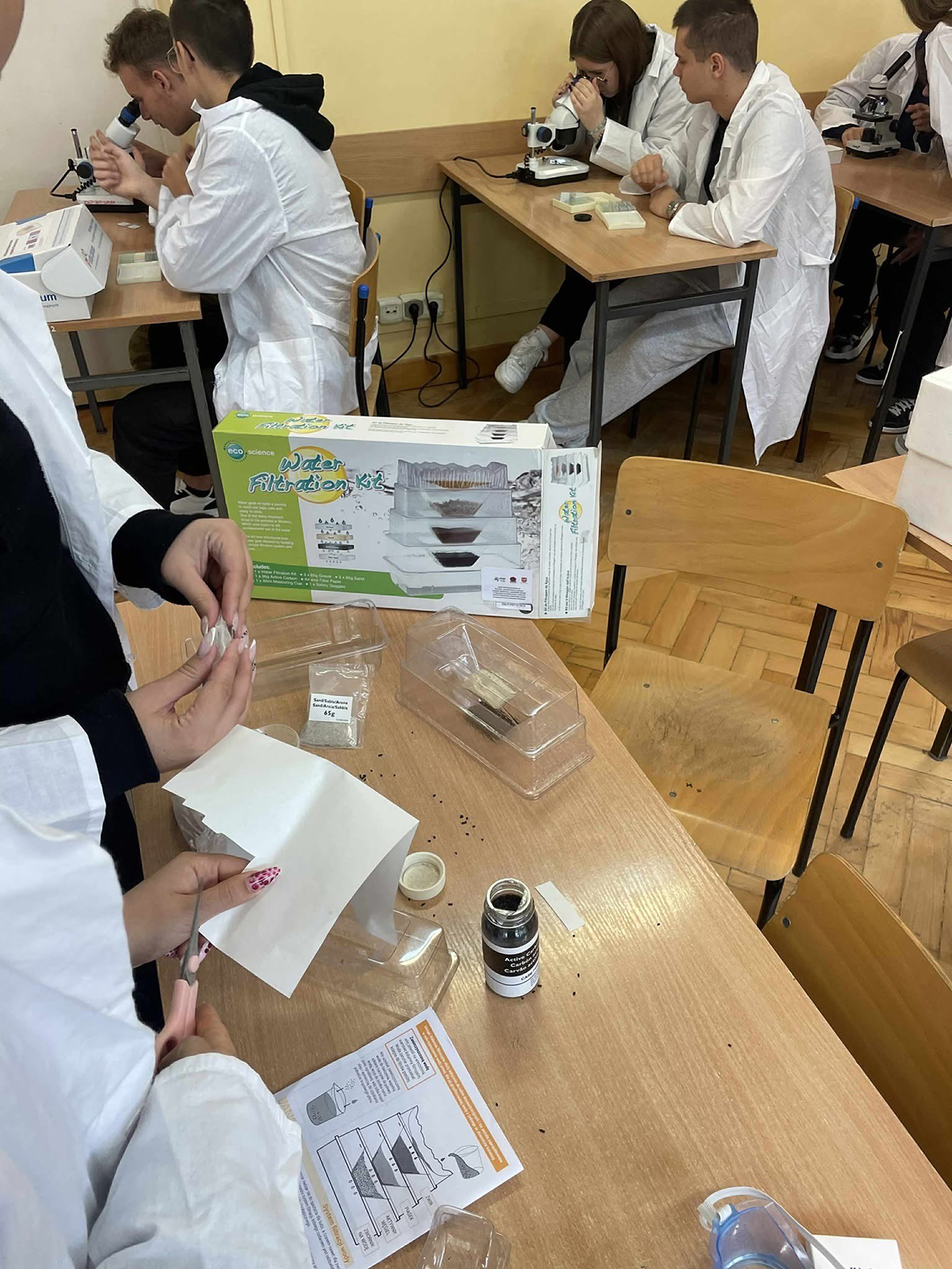 Grupa dzieci ubranych w białe fartuchy stoi przy stole analizując dane lub wyniki eksperymentu. Są skupione i zaangażowane w wykonywane zadanie