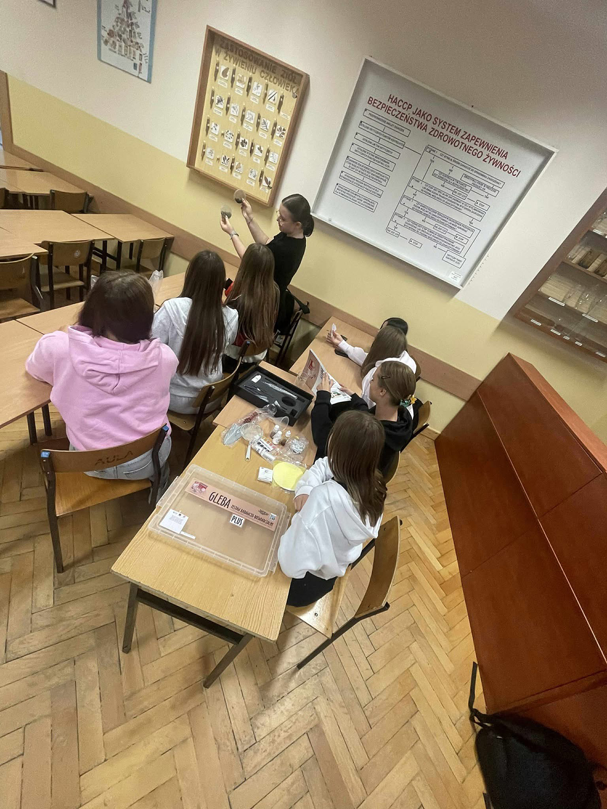 Zdjęcie z lotu ptaka przedstawiające sześciu uczniów siedzących w klasie. Dzieci mają na sobie kolorowe ubrania i siedzą przy stołach zajęte pracą lub rozmową. Wokół nich widoczne są szkolne pomoce i materiały edukac