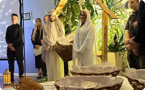 Na zdjęciu widać scenę z przedstawienia bożonarodzeniowego. W centrum stoi drewniana konstrukcja symbolizująca stajenkę, pod którą znajdują się dwie osoby ubrane na biało odgrywające role Maryi i Józefa.