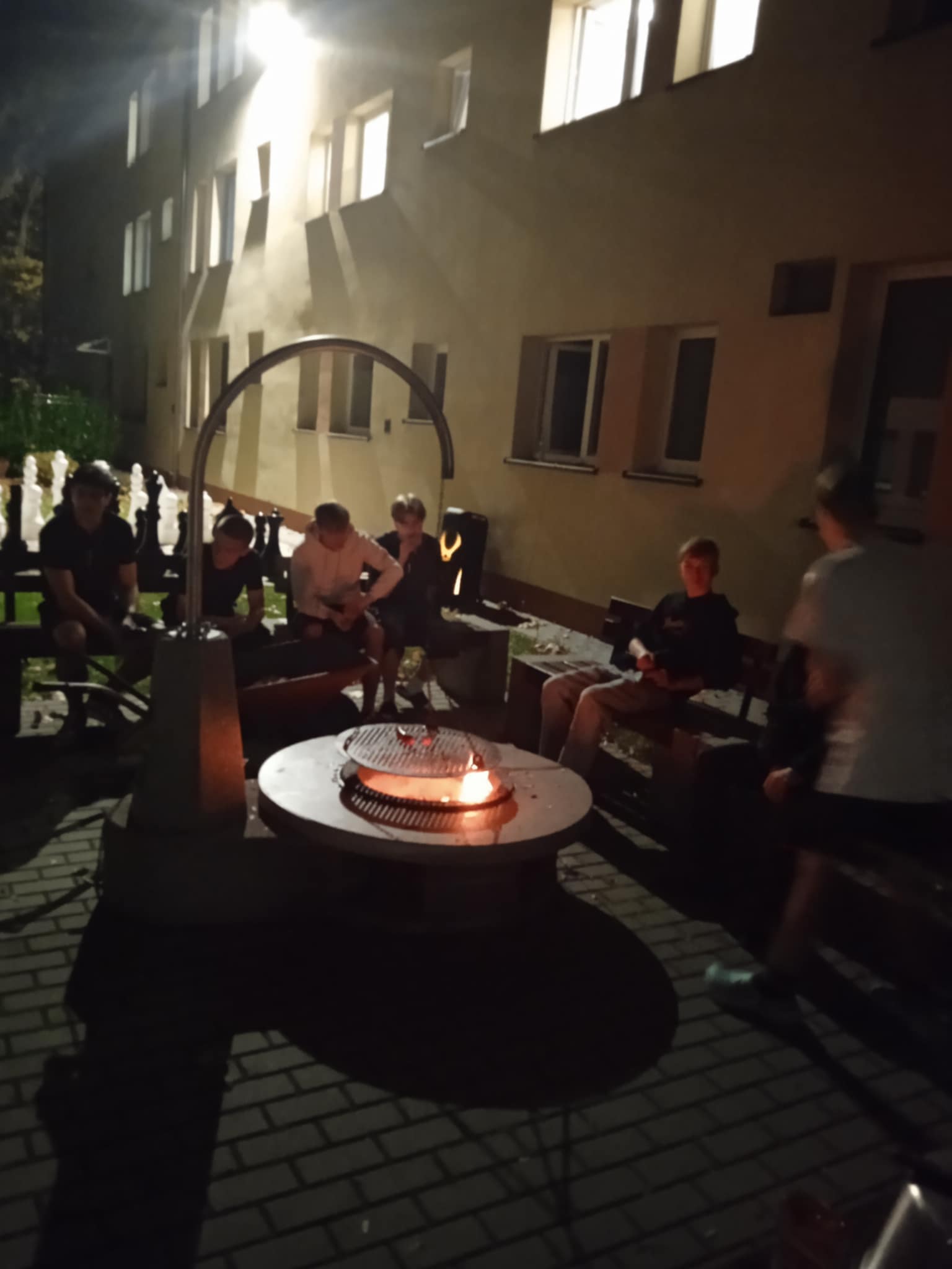 Grupa młodych osób siedzi wieczorem przy palenisku na dziedzińcu przy budynku internatu. Jest ciemno, a scena oświetlona jest światłem z okien i lekkim blaskiem ognia. Ludzie siedzą na ławkach w półokręgu, rozmawiają i odpoczywają.