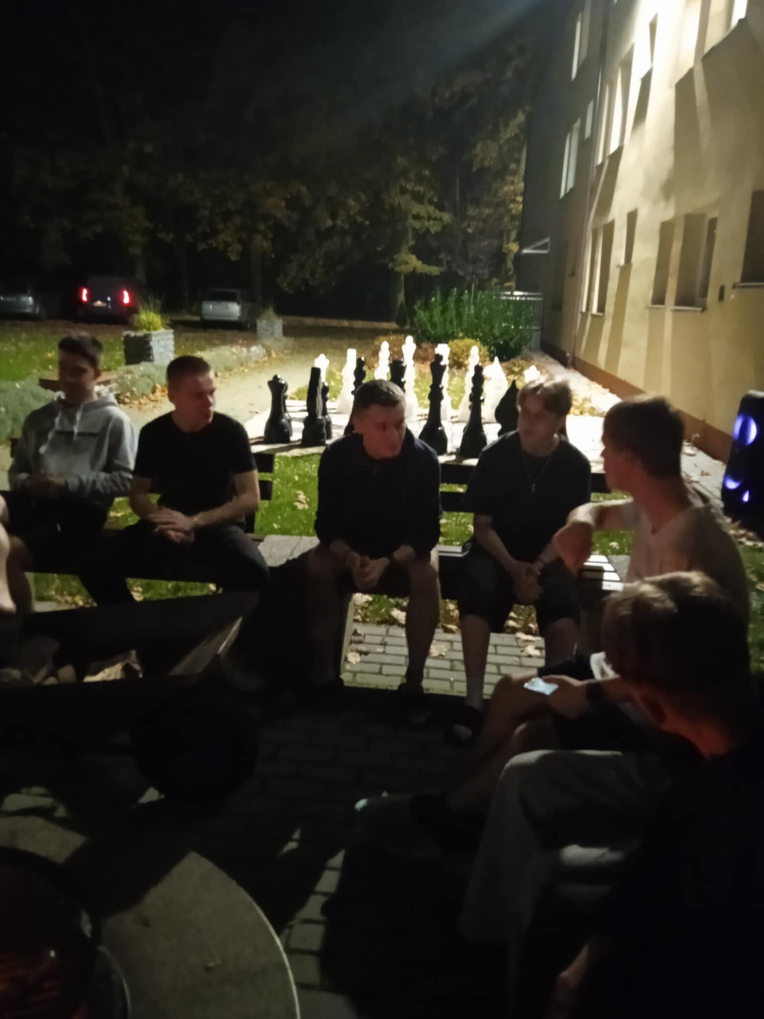Kilku młodych chłopaków siedzi na ławce na zewnątrz budynku. W tle widać duże figury do szachów ustawione na trawniku. Jest noc, a jedyne światło pochodzi z lamp i okien budynku. Osoby wyglądają na skupione na rozmowie.