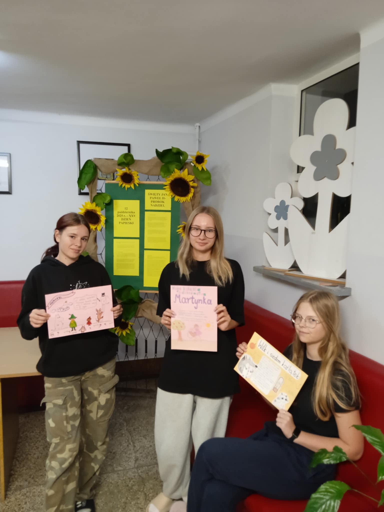 Trzy dziewczyny stoją w szkolnym korytarzu. Każda trzyma w rękach kolorowy plakat przedstawiający wybraną książkę dla dzieci. W tle widać tablicę ozdobioną słonecznikami.