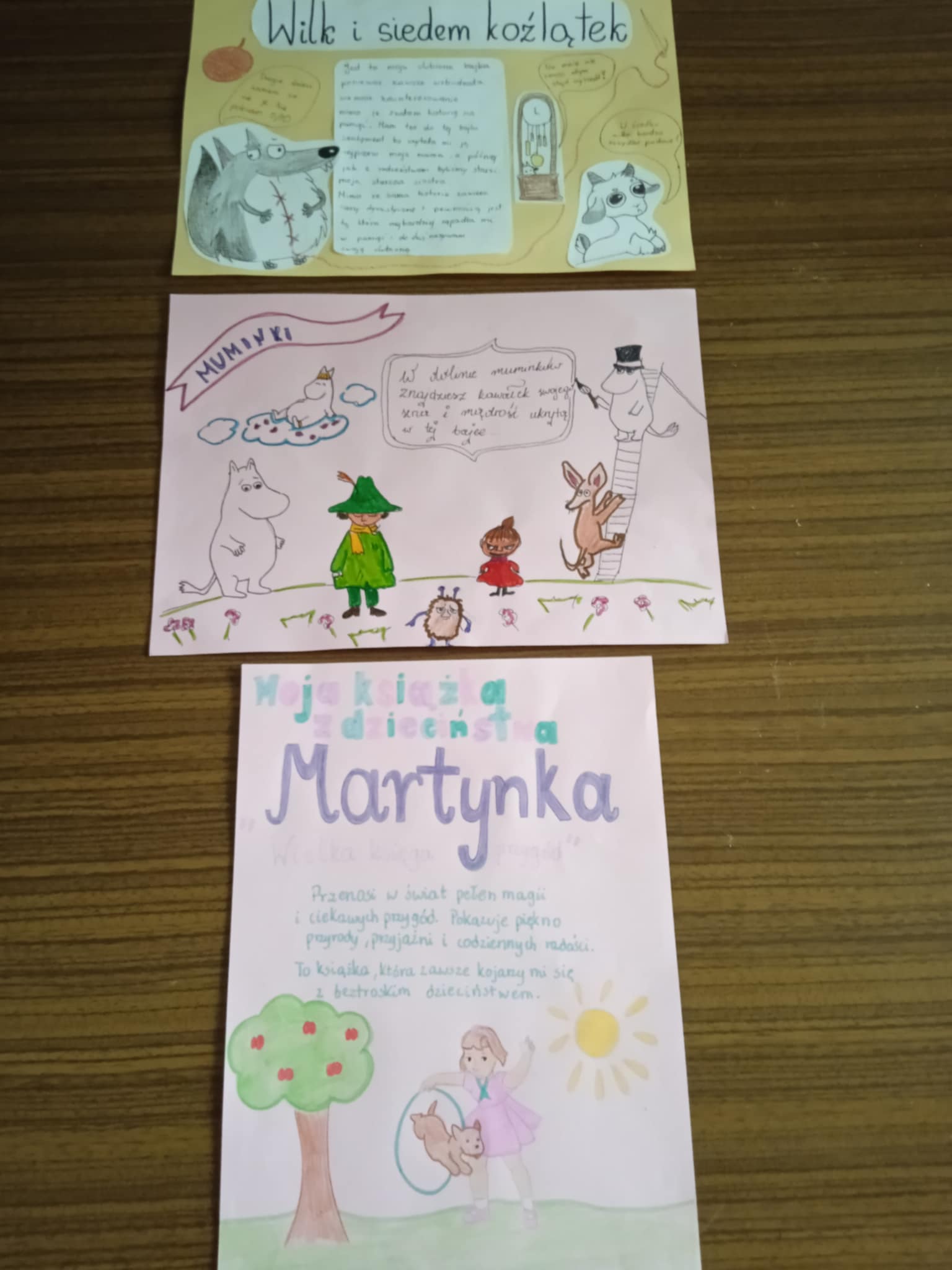 Na stole leżą trzy kolorowe plakaty wykonane przez uczniów. Przedstawiają: „Wilk i siedem koźlątek”, „Muminki” oraz książkę „Martynka”. Plakaty są ozdobione rysunkami i krótkimi opisami.