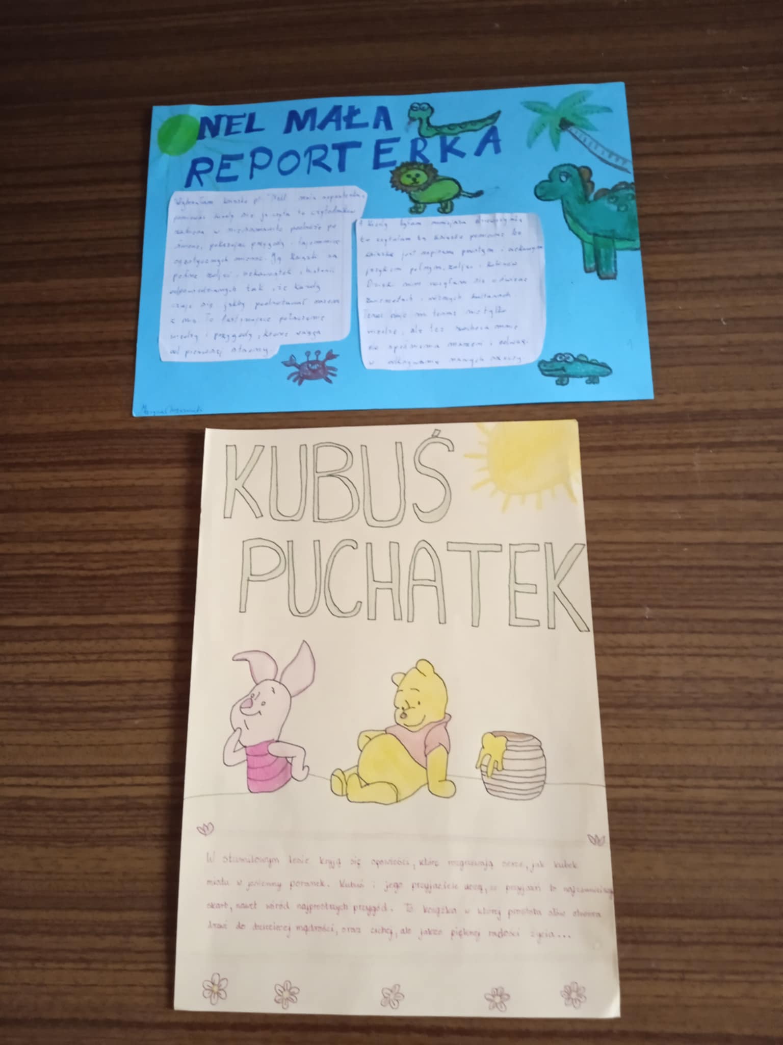 Na stole leżą dwa kolejne plakaty: „Nel – mała reporterka” w kolorze niebieskim z rysunkami zwierząt oraz „Kubuś Puchatek” w kolorze jasnym, z rysunkami misia i prosiaczka. Plakaty są starannie wykonane i kolorowe.