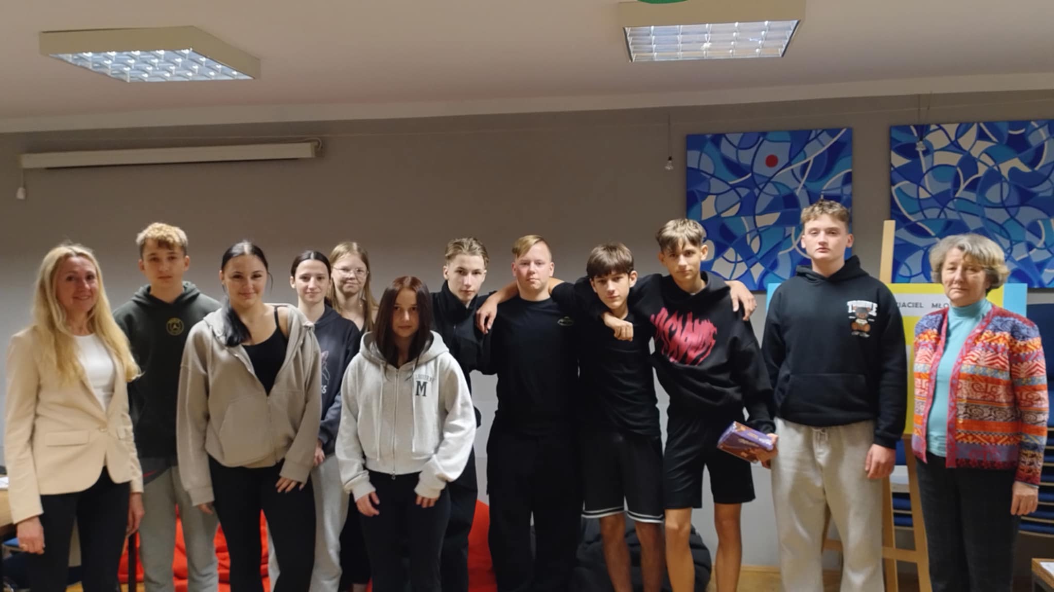 Grupa młodych osób – uczestników konkursu o Patronie Szkoły stoi razem w sali, pozując do wspólnej fotografii. Towarzyszą im dwie dorosłe osoby. W tle widoczne są kolorowe obrazy i elementy wyposażenia sali.