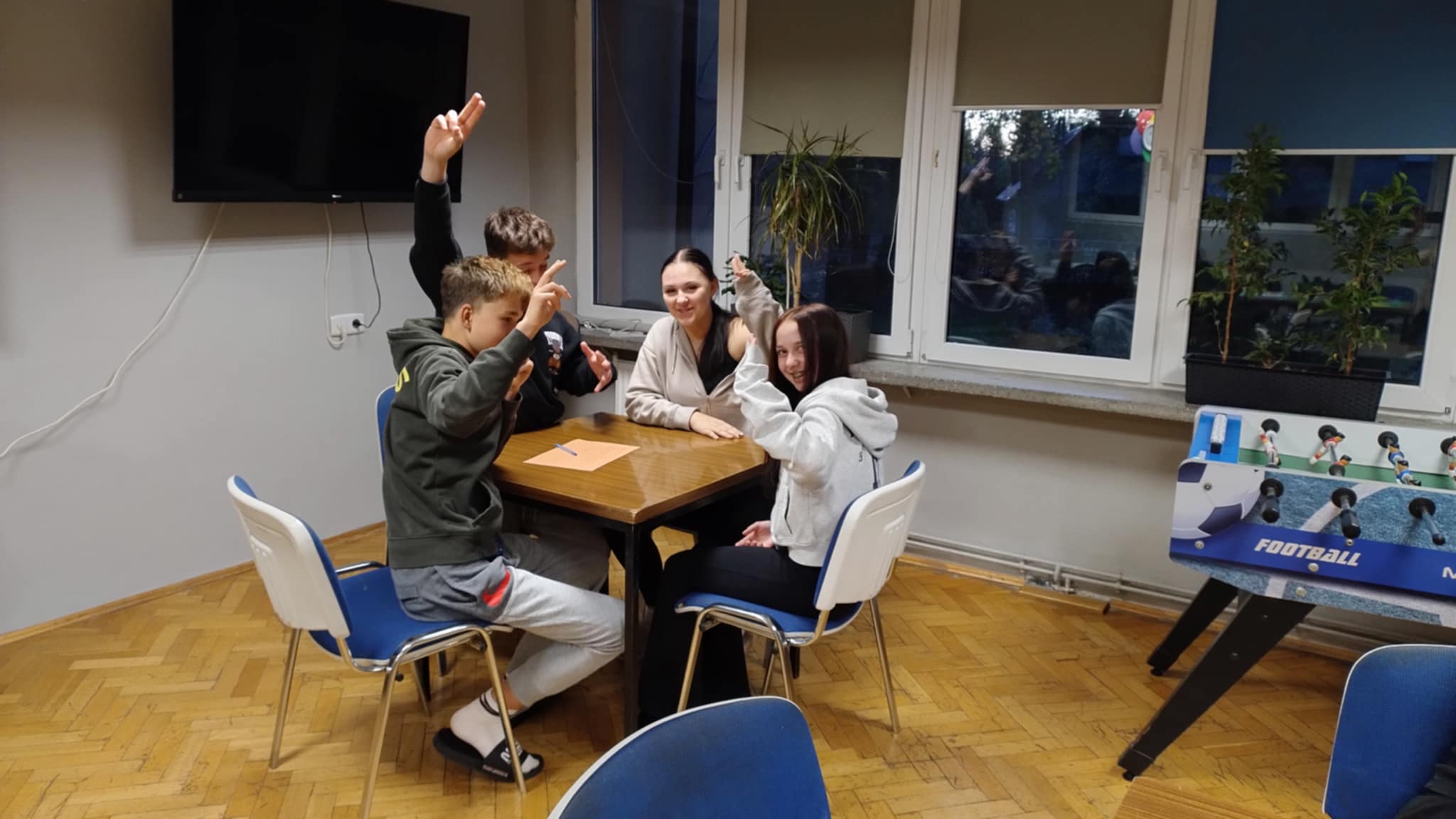 Kilka osób siedzi przy stole w świetlicy internatu. Wszyscy wyglądają na zaangażowanych i uśmiechniętych, unoszą ręce, prawdopodobnie zgłaszając się do udziału w proponowanych aktywnościach, co tworzy swobodną i radosną atmosferę.