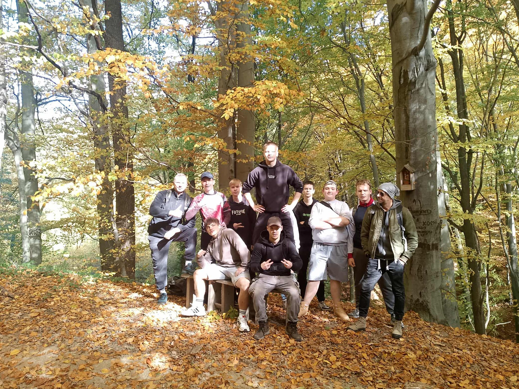 Grupa młodych osób oraz dorosły stoi razem w lesie. Jest słoneczny dzień, a na ziemi leżą żółte jesienne liście. Wszyscy pozują do zdjęcia, niektórzy siedzą na ławce, inni stoją obok drzew.