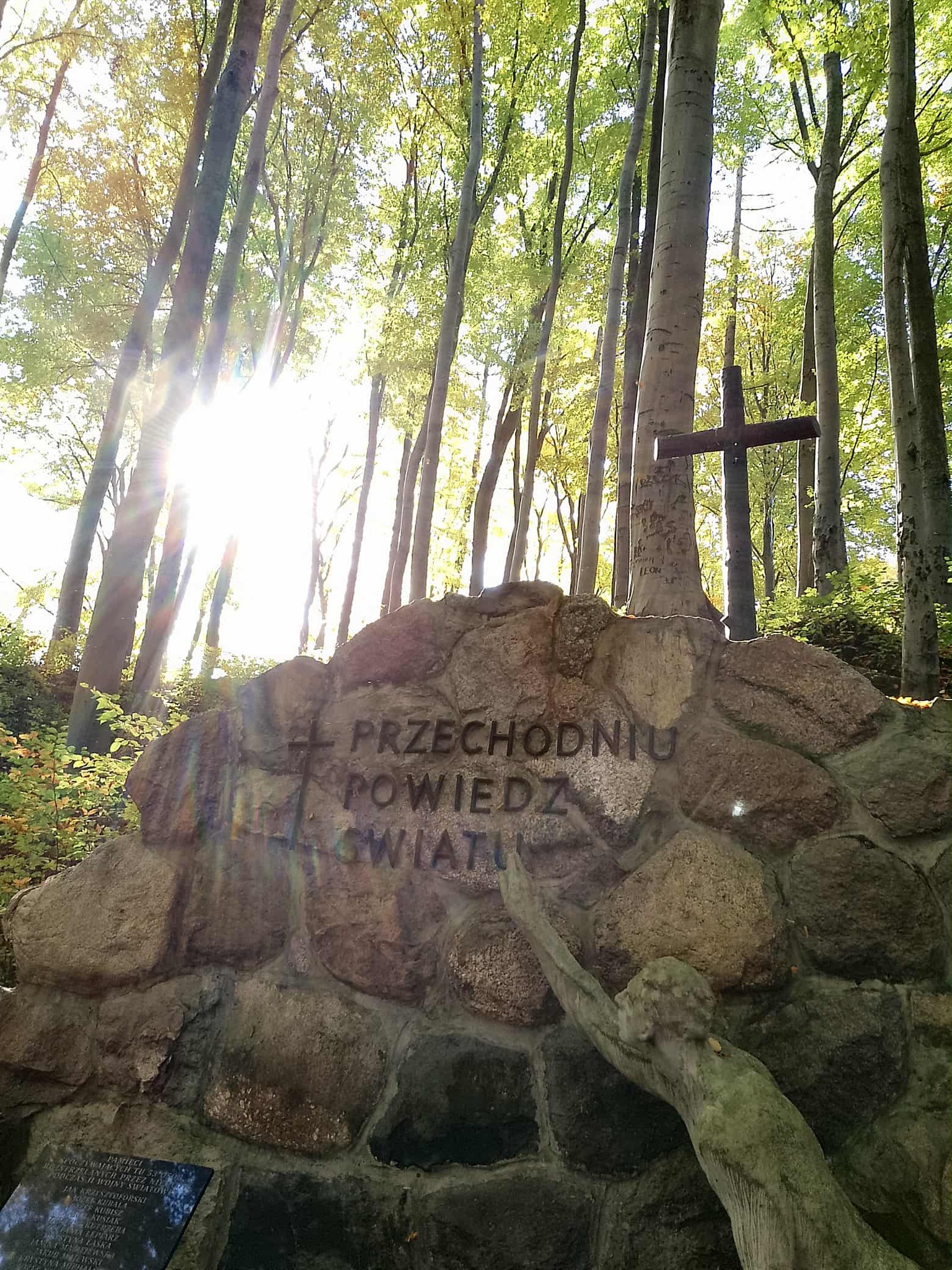 W lesie stoi kamienny pomnik z krzyżem i napisem „Przechodniu, powiedz światu…”. Obok kamieni znajduje się rzeźba postaci wyciągającej rękę ku górze. Przez drzewa przebija mocne światło słoneczne, tworząc jasne promienie.