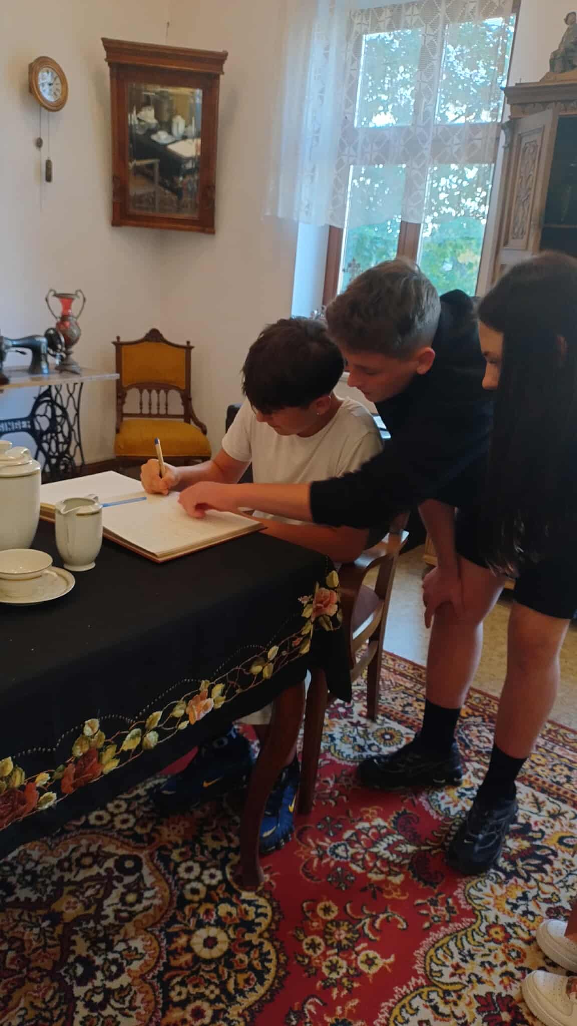 3.1	W pomieszczeniu Izby regionalnej w Wojniczu troje młodych osób siedzi i stoi przy stole. Jedna osoba pisze w dużej księdze, a dwie pozostałe pomagają jej lub przyglądają się. Wokół znajdują się stare meble, ozdobny dywan oraz elementy dekoracyjne, co nadaje miejscu muzealny lub historyczny charakter.