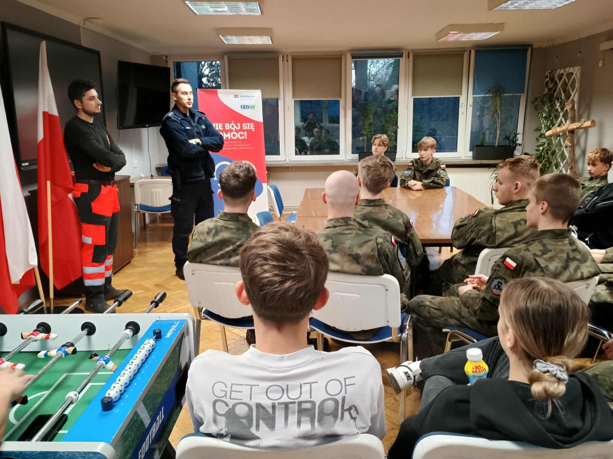 2.	Grupa młodzieży, głównie w mundurach, siedzi na krzesłach i słucha dwóch prowadzących: jednego w policyjnym mundurze, drugiego w stroju ratownika. W tle widoczny jest baner promujący projekt „Nie bój się pomóc!”.