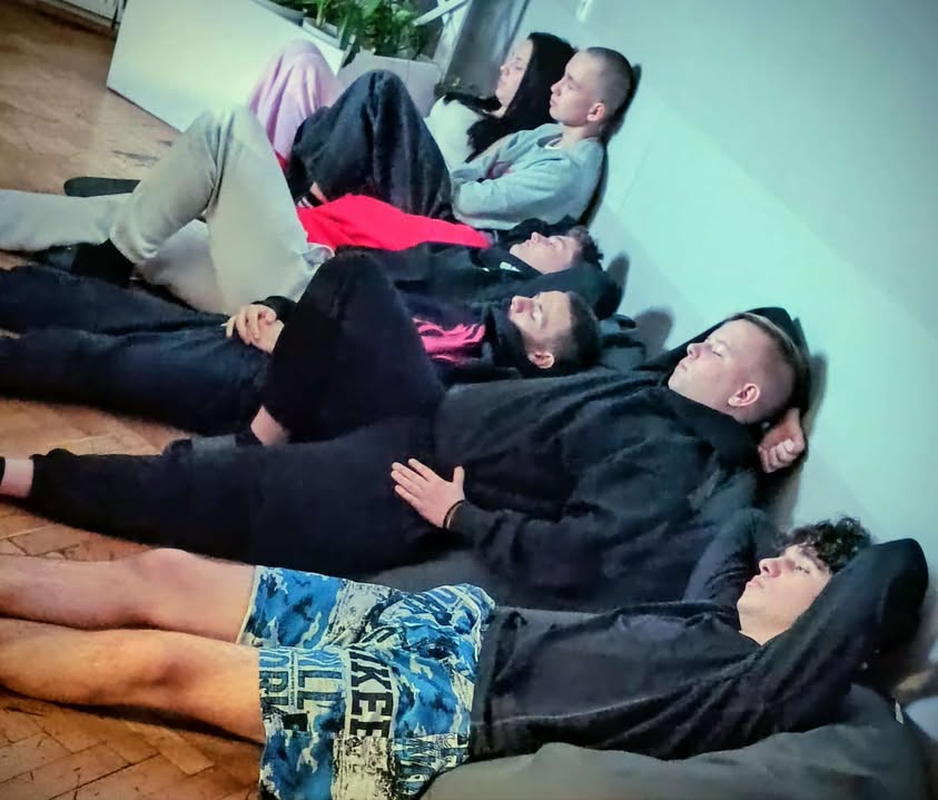 Kilka osób leży wygodnie na podłodze na matach lub kocach. Mają zamknięte oczy i odpoczywają, jak podczas ćwiczeń wyciszających lub relaksacji.