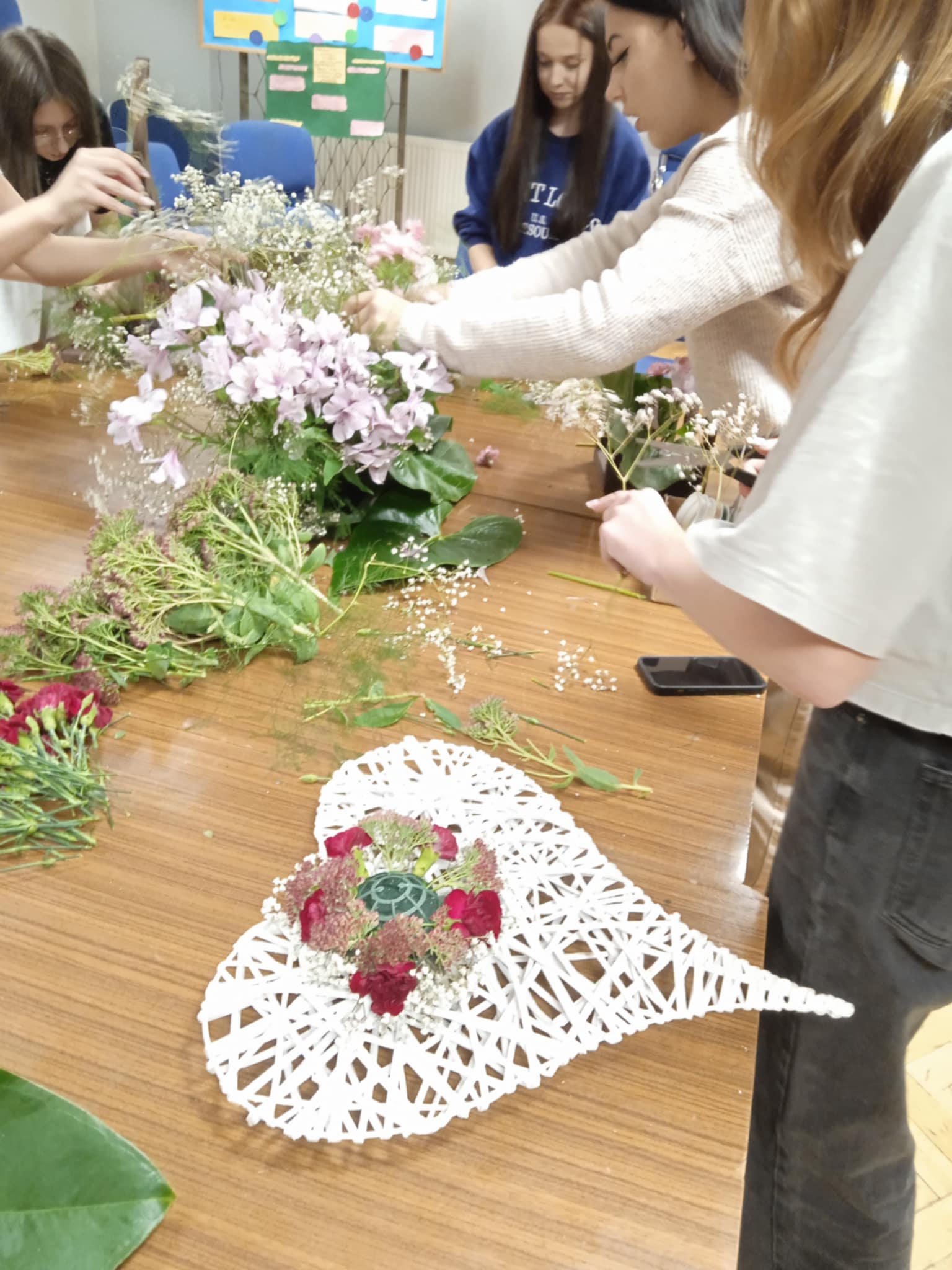 Kilka osób stoi przy dużym stole i wspólnie układa kwiaty. Na blacie leżą różne rośliny i białe serce z wikliny, w którym powstaje kwiatowa dekoracja. Wszyscy skupiają się na pracy.