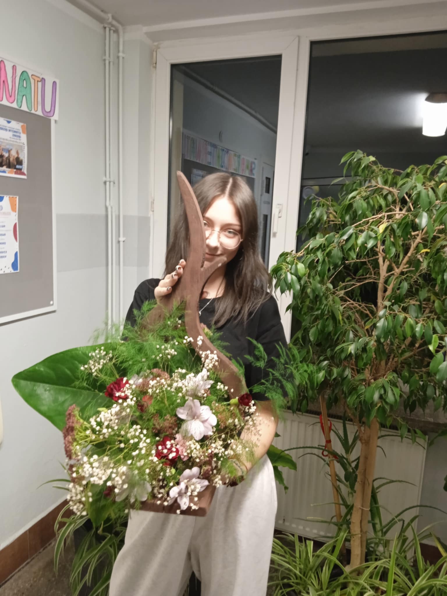 Młoda osoba stoi na korytarzu i trzyma w rękach dużą kompozycję kwiatową. W koszu znajdują się zielone liście, białe i różowe drobne kwiaty oraz czerwone akcenty. W tle widać rośliny doniczkowe i drzwi.