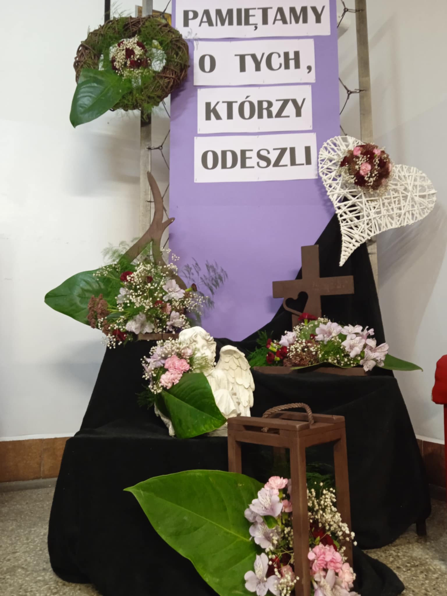 Na korytarzu ustawiono dekoracyjną wystawę z okazji pamięci o zmarłych. Na czarnym materiale stoją kompozycje kwiatowe, figurka anioła, latarenka i duże zielone liście. W tle wiszą tabliczki z napisem: „PAMIĘTAMY O TYCH, KTÓRZY ODESZLI”.