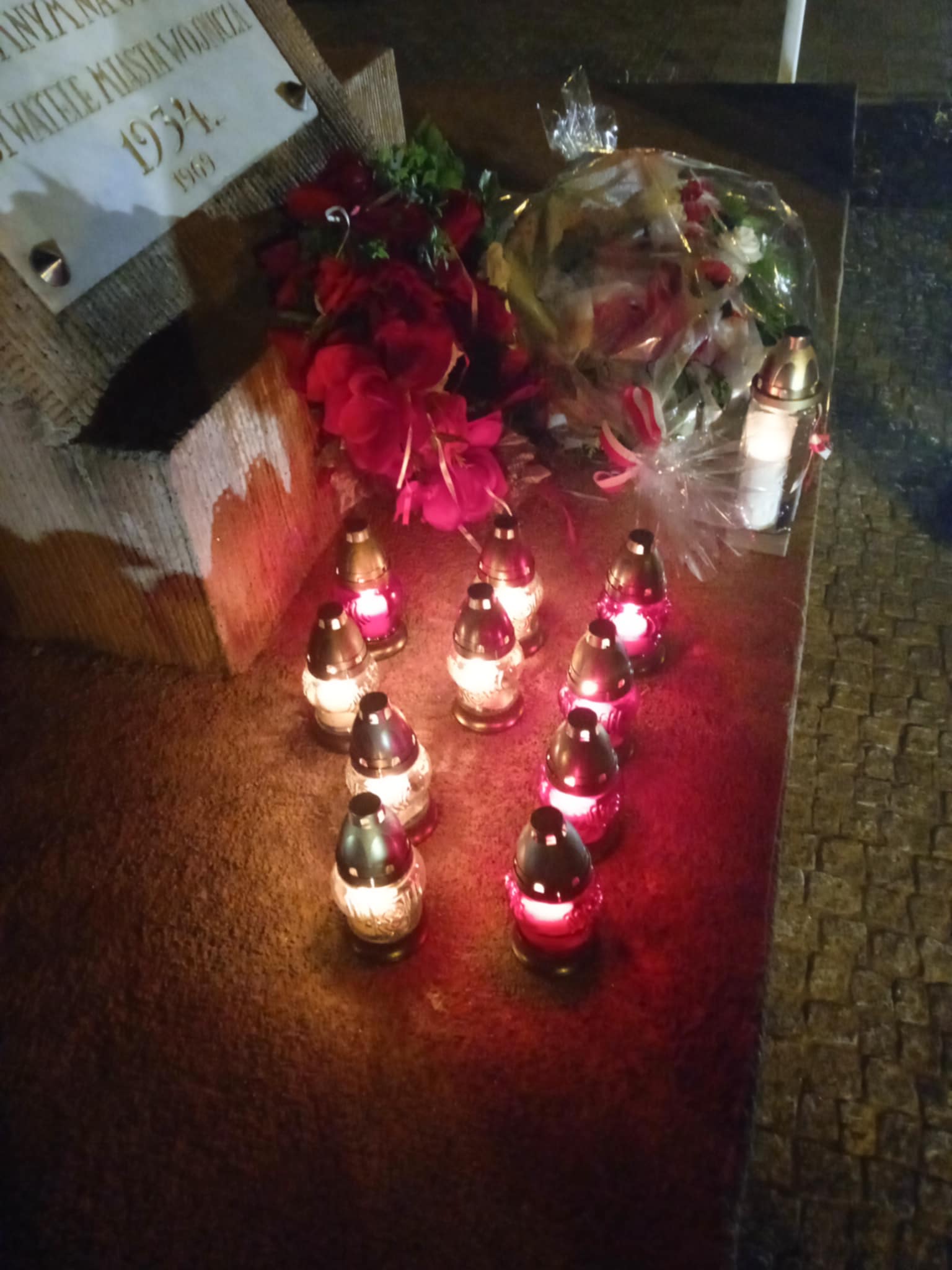 Na pomniku lub płycie pamiątkowej leżą czerwone kwiaty i biało-czerwony wieniec. Przed pomnikiem stoi kilka zapalonych lampek – czerwonych i białych – ustawionych w rządkach.