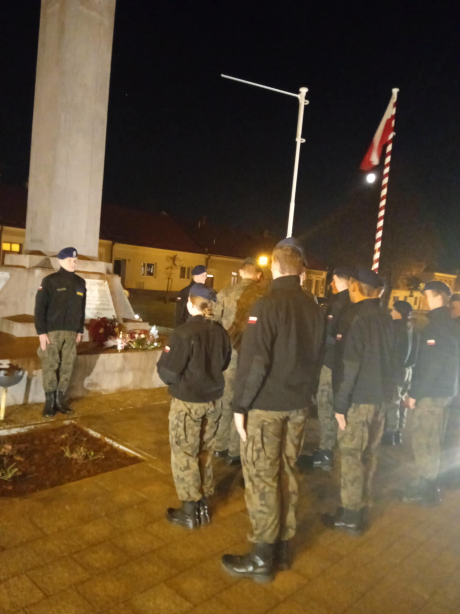 . Kilka osób w mundurach stoi w szeregu przy wysokim pomniku. Przed pomnikiem widoczne są kwiaty i zapalone znicze. Obok pomnika powiewa biało-czerwona flaga. Jest noc, a miejsce oświetlają latarnie.