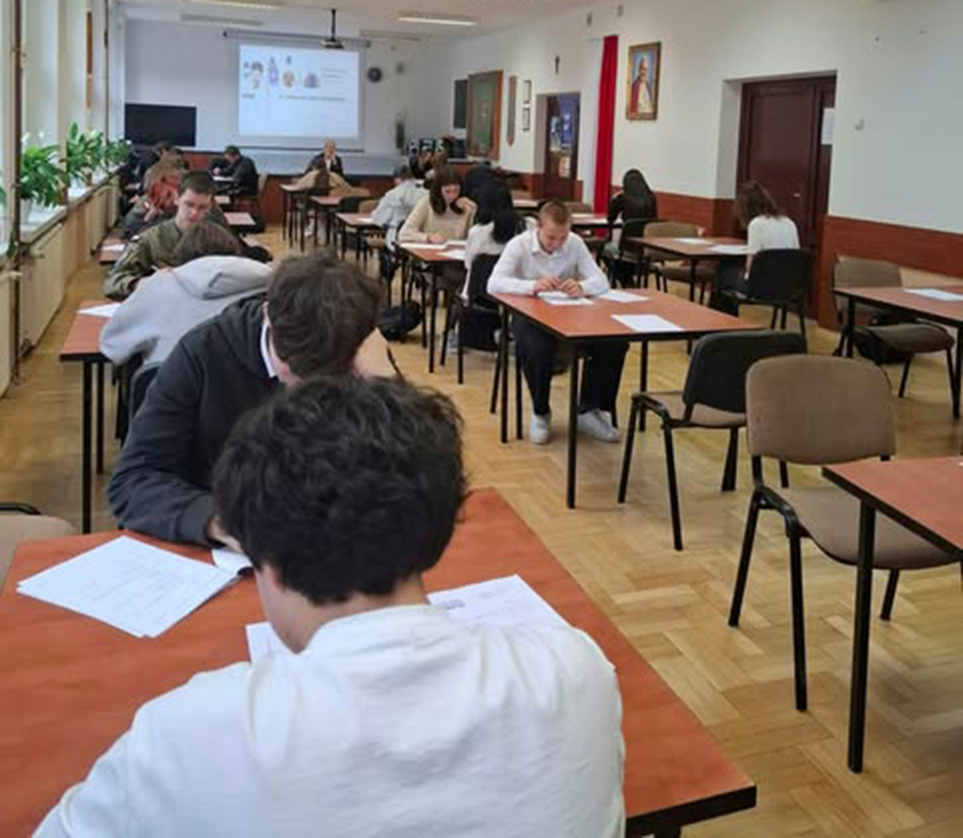 Zdjęcie przedstawia uczniów i uczennice piszące w sali test podczas pierwszego etapu szkolnego Olimpiady Języka Angielskiego. W tle widać wyświetlone logo Olimpiady.