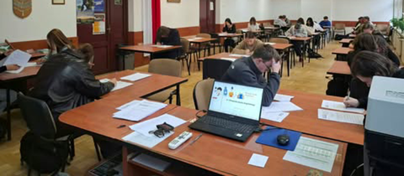 Zdjęcie przedstawia uczniów i uczennice piszące w sali test z innej perspektywy. Widać uczniów i uczennice skupionych nad testem.